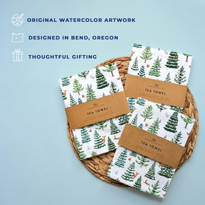Set of tea towels with forest design on a woven tray against a light blue background.