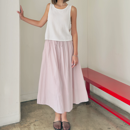 Person wearing a white top and pink skirt standing against a plain wall with a red bench.