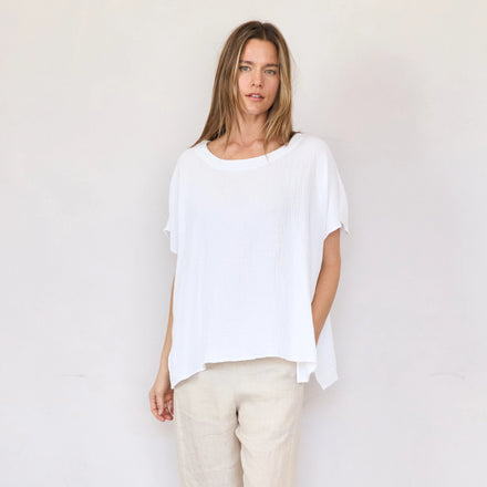 Woman wearing a white top and beige pants standing against a plain white wall.