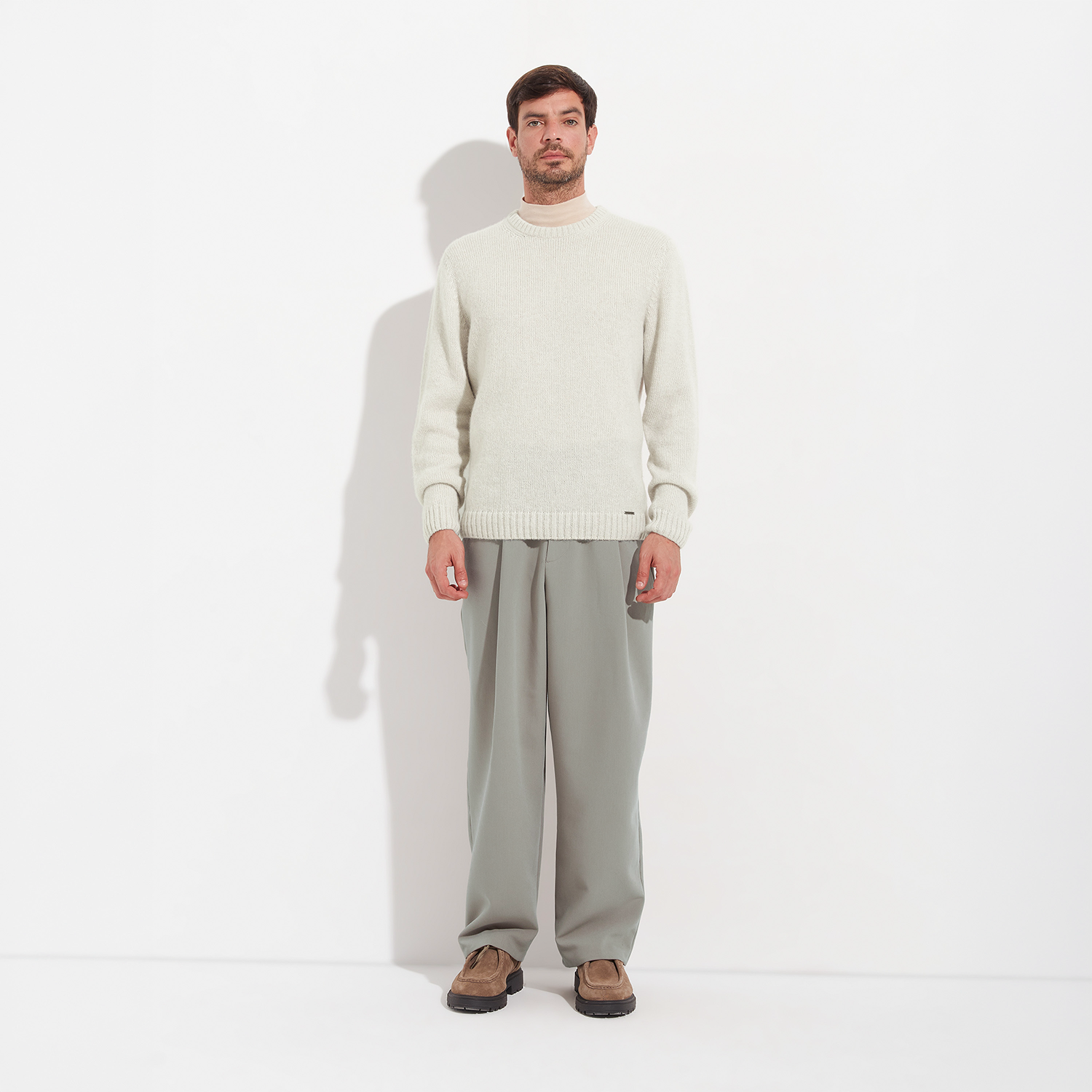 Man wearing a cream sweater and gray pants standing against a white background