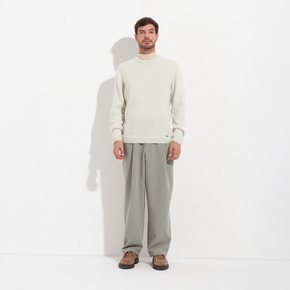 Man wearing a cream sweater and gray pants standing against a white background