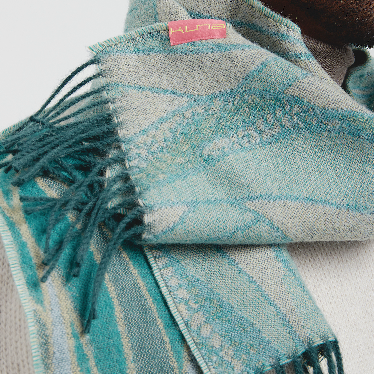 Teal and gray patterned scarf with fringes and a visible brand label on a light background