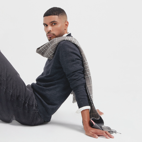 Man sitting on a white floor wearing a dark sweater, plaid scarf, and gray pants.