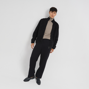 Man wearing a dark grey quilted jacket, beige turtleneck, and black pants on a white background