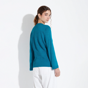 Woman wearing a teal sweater and white pants on a light gray background