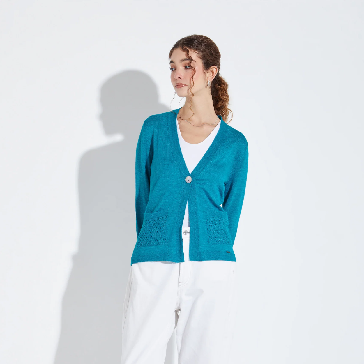 Woman wearing a teal cardigan over a white shirt and pants on a light gray background