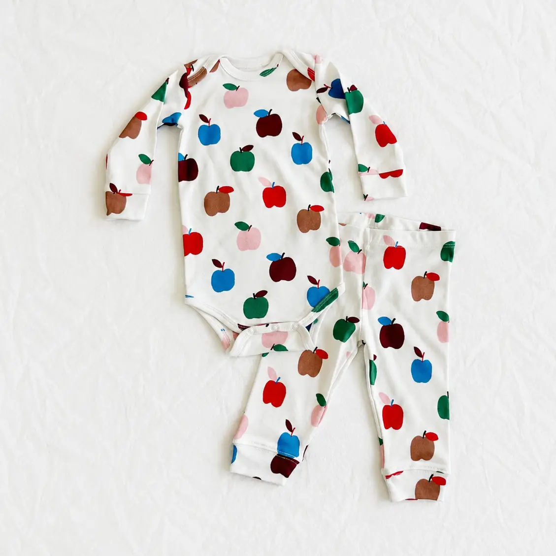 A baby long-sleeve bodysuit and matching pants set laid flat on a white textured background. The set features a white base with a colorful pattern of stylized apples in red, green, blue, brown, and pink.