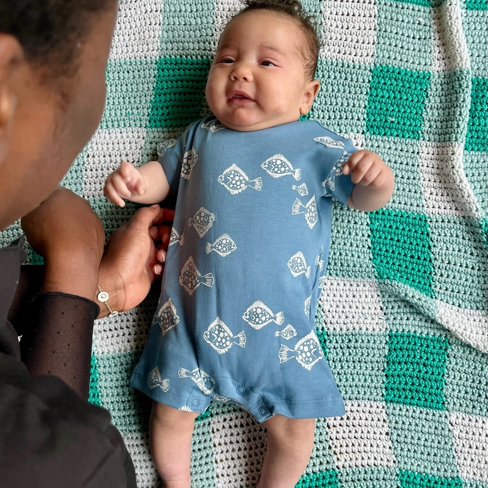 Baby in a blue onesie with white patterns lying on a green and white checkered blanket, being held by an adult.