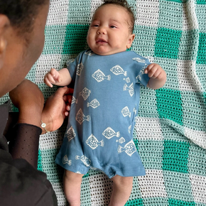 Baby in a blue onesie with white patterns lying on a green and white checkered blanket, being held by an adult.