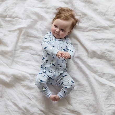 Baby wearing a blue onesie with bird patterns lying on a white bed.
