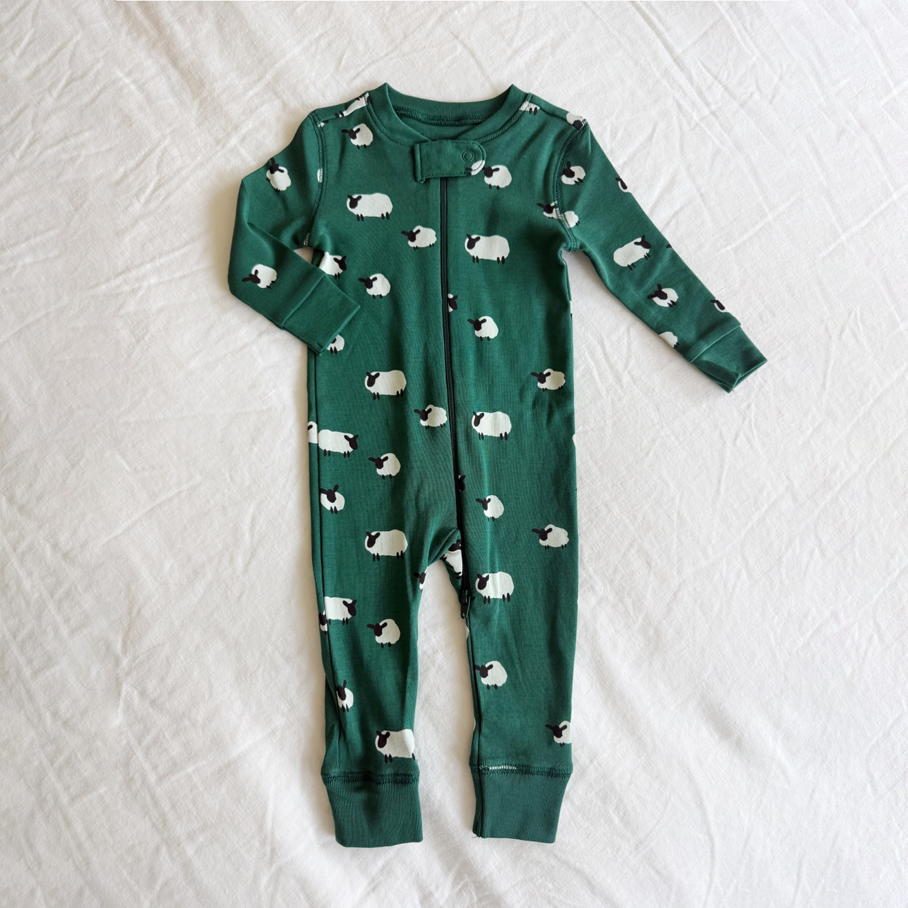 A baby long-sleeve footless one-piece pajama in dark green with a repeating pattern of white sheep with black faces, laid flat on a white textured fabric background.