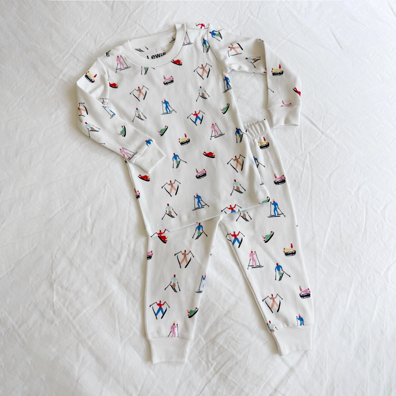 A two-piece long-sleeve pajama set laid flat on a white fabric background. The set is white with a colorful all-over print of skiers and snowboarders.