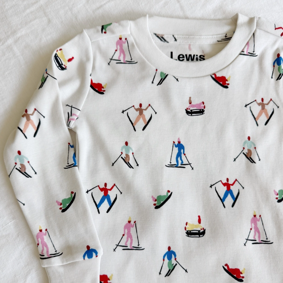 A white long-sleeve baby pajama top with an all-over print of skiers in colorful outfits, laid flat on a white textured fabric background. The brand name 'Lewis' is printed inside the collar.