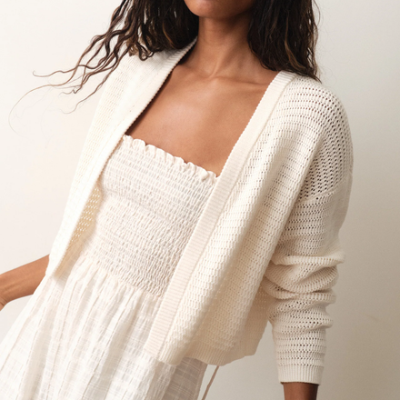 Woman wearing a white crochet cardigan over a white dress against a neutral background