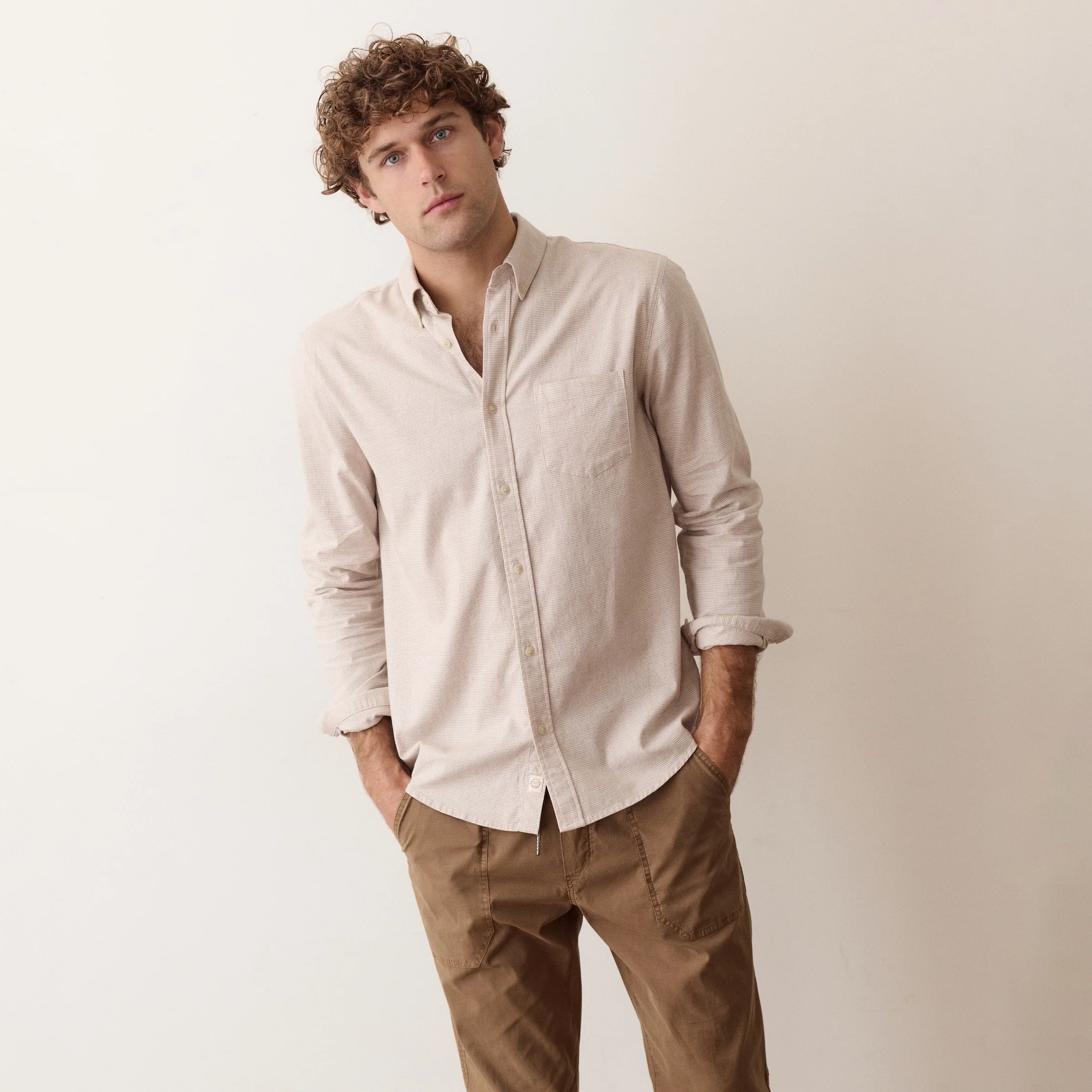 A man wearing a light beige long-sleeve linen button-down shirt and brown trousers, standing against a plain off-white background.