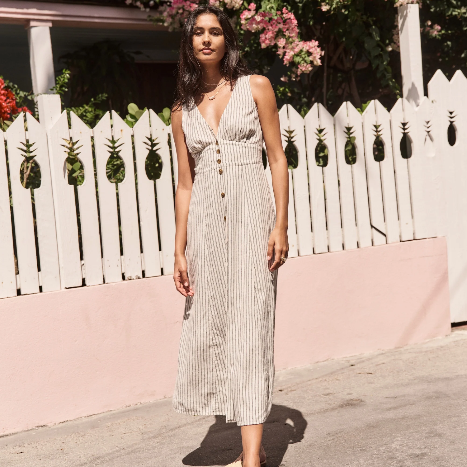 Woman in a long dress standing in front of a white picket fence with pineapple designs.