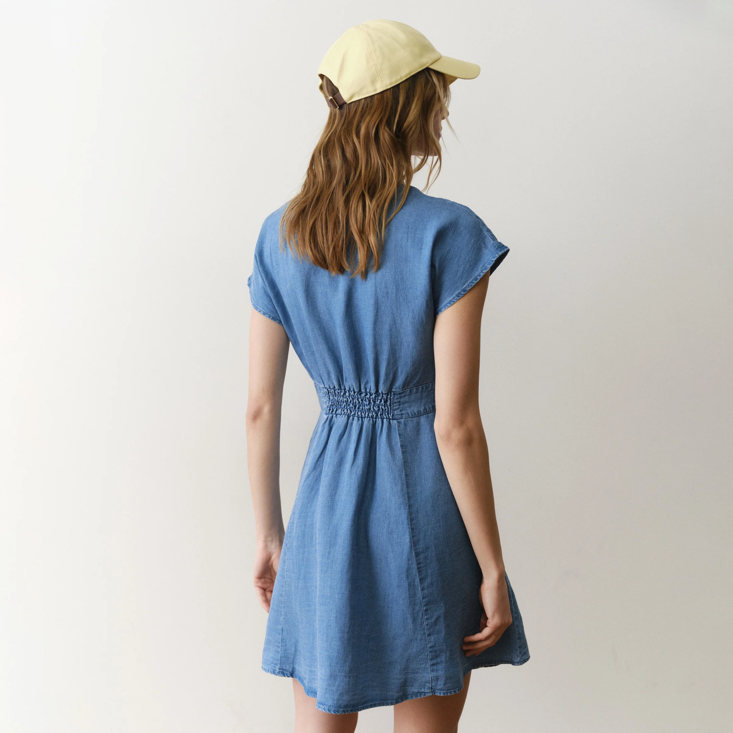 Woman wearing a blue dress and yellow cap from behind on a plain background
