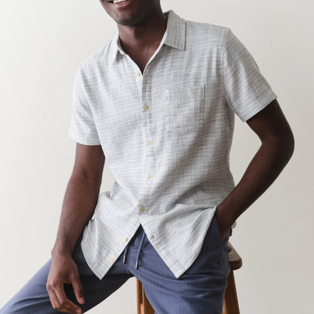 Man wearing a light blue short-sleeve shirt with a subtle pattern, sitting on a wooden stool against a plain background.