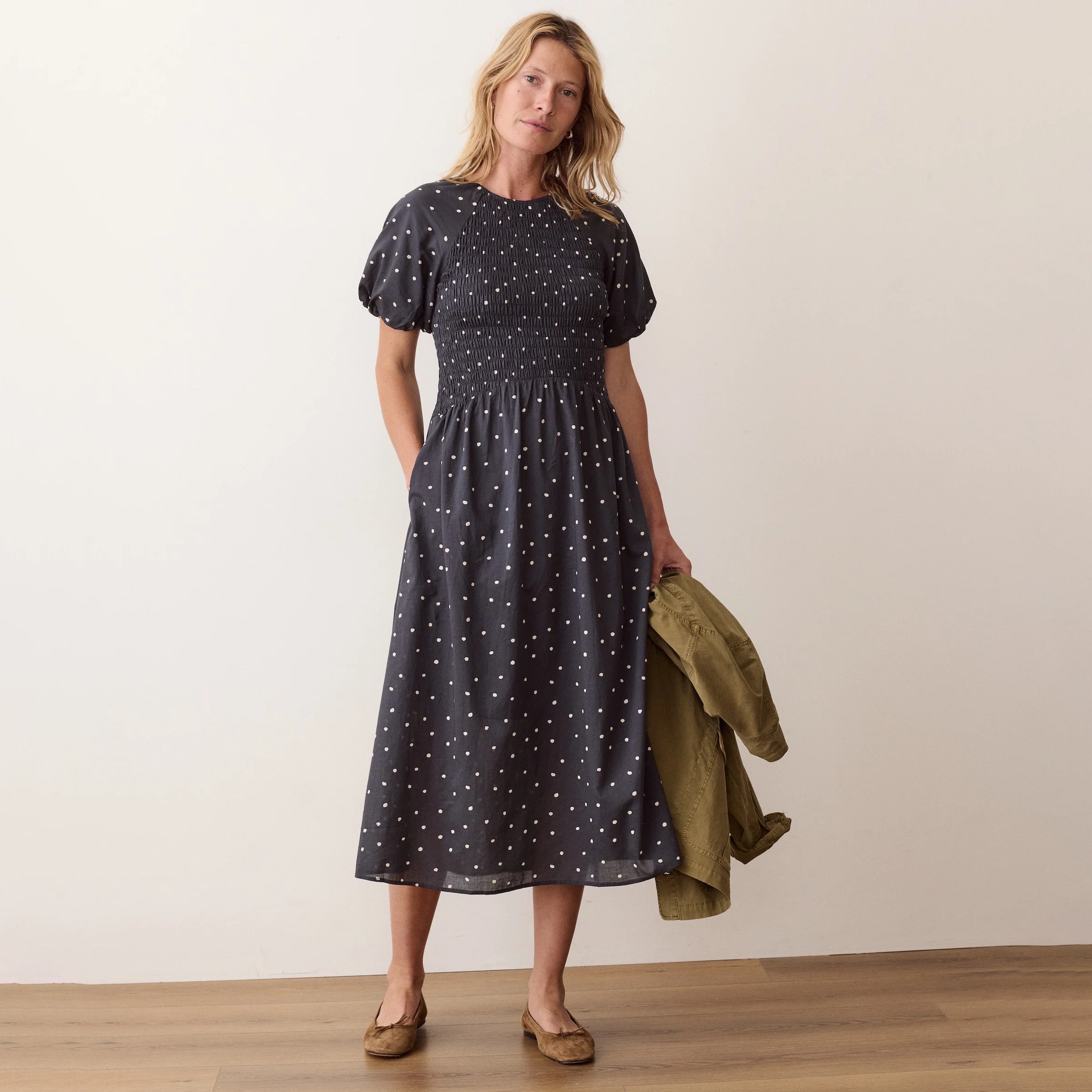 A woman stands in a studio wearing a dark navy midi dress with white polka dots, holding an olive green jacket. The background is a clean off-white wall with a light wood floor.