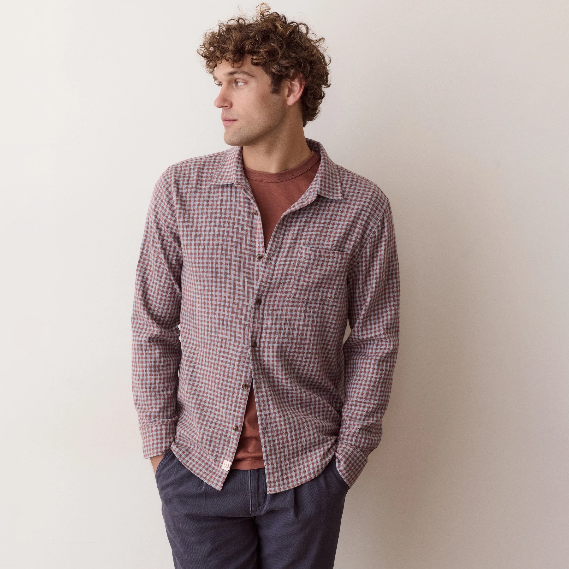 A man wearing a checkered long-sleeve button-down shirt over a rust-colored t-shirt and dark gray pants, standing against a plain off-white wall.