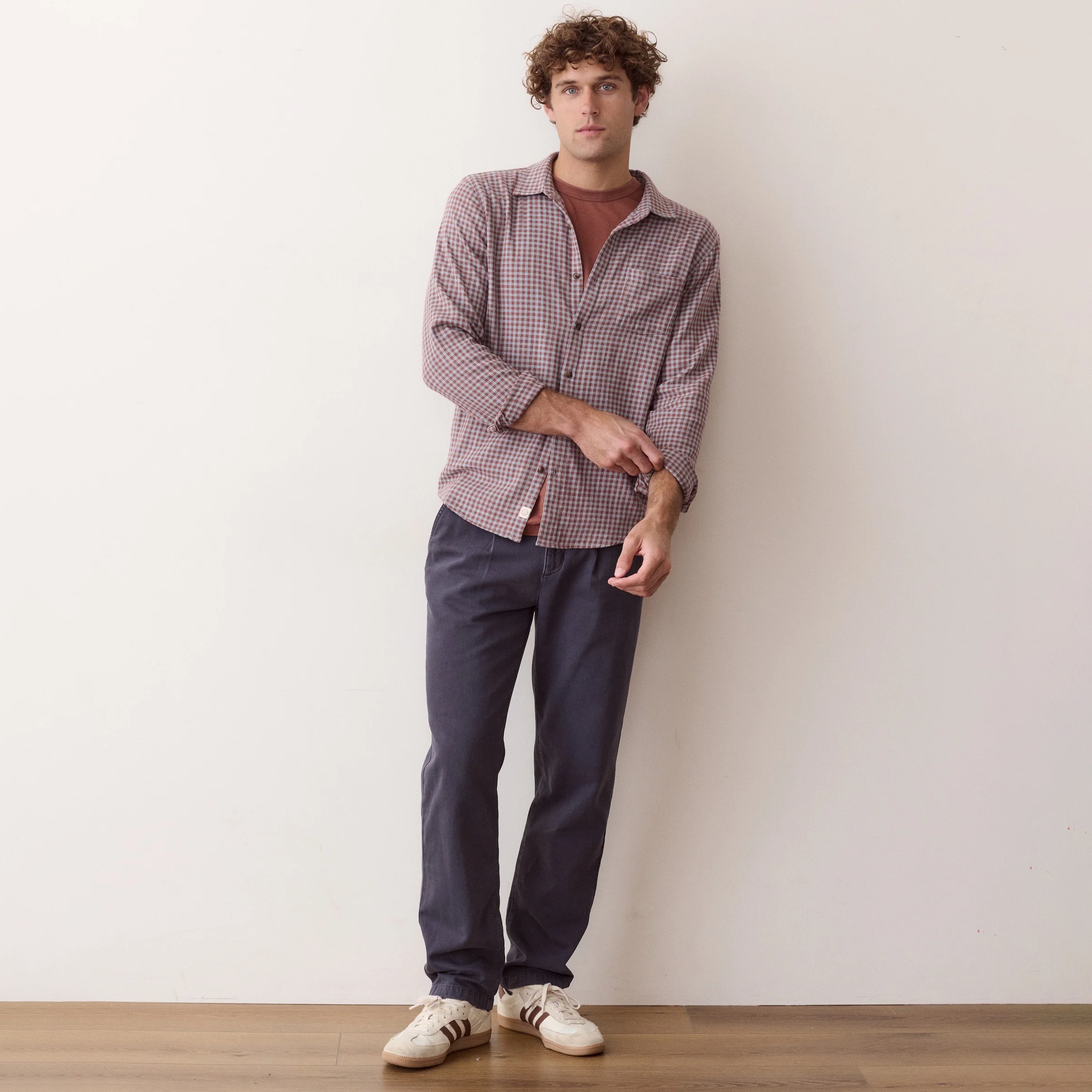 A man stands against a plain off-white wall wearing a long-sleeve button-up shirt with a fine red gingham pattern, dark charcoal gray trousers, and white sneakers with black stripes.