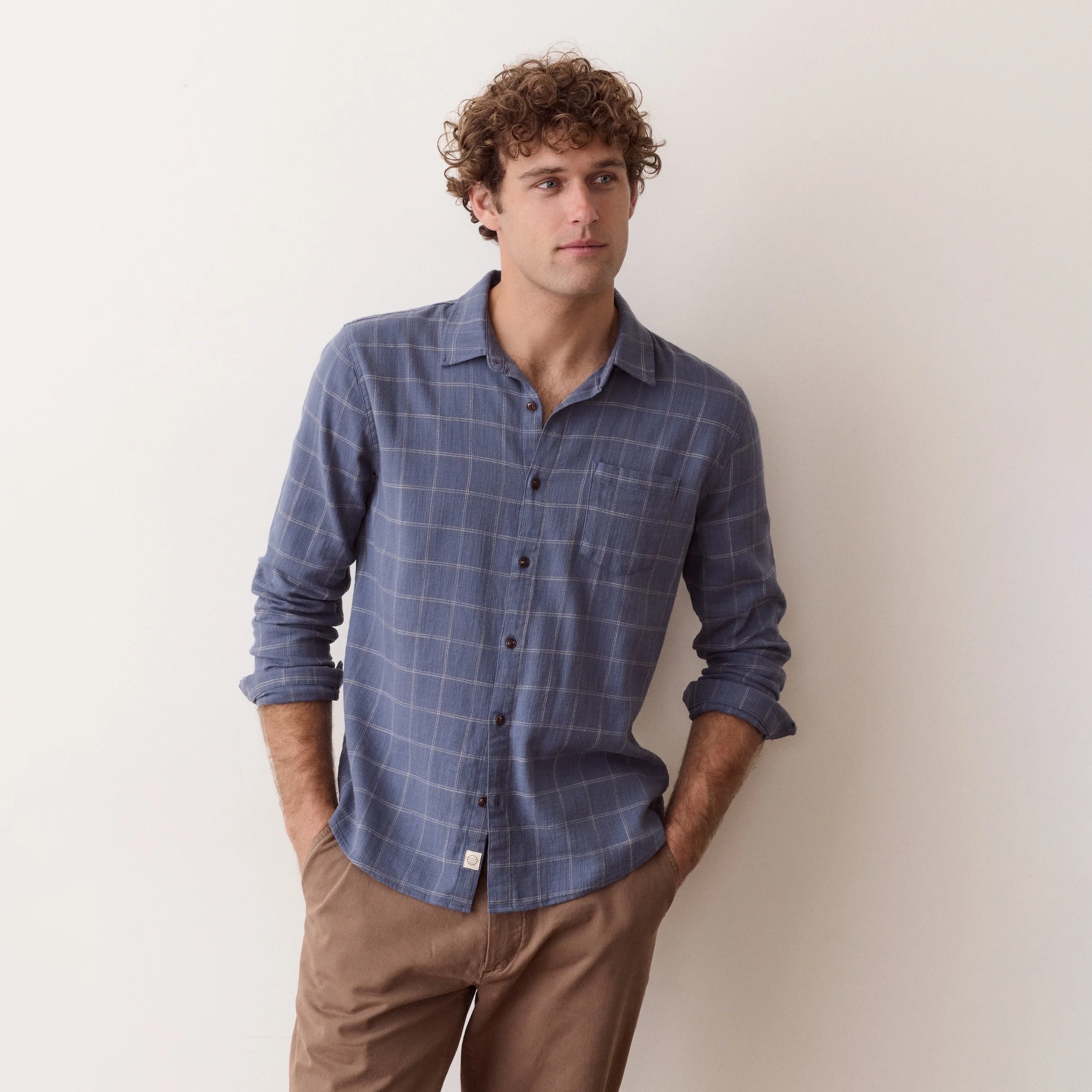 A man with curly brown hair wearing a blue and gray plaid button-down shirt and brown pants, standing against a plain off-white wall.