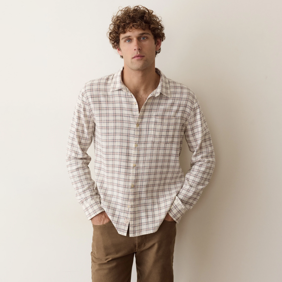 A man with curly brown hair wearing a beige and brown plaid button-down shirt and brown pants, standing against a plain off-white wall.
