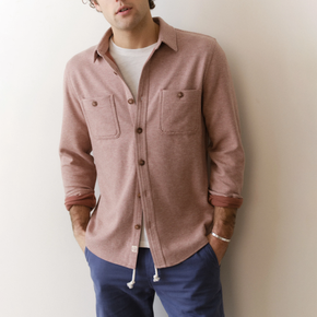 Man wearing a pink button-up shirt over a white t-shirt with blue pants against a plain background