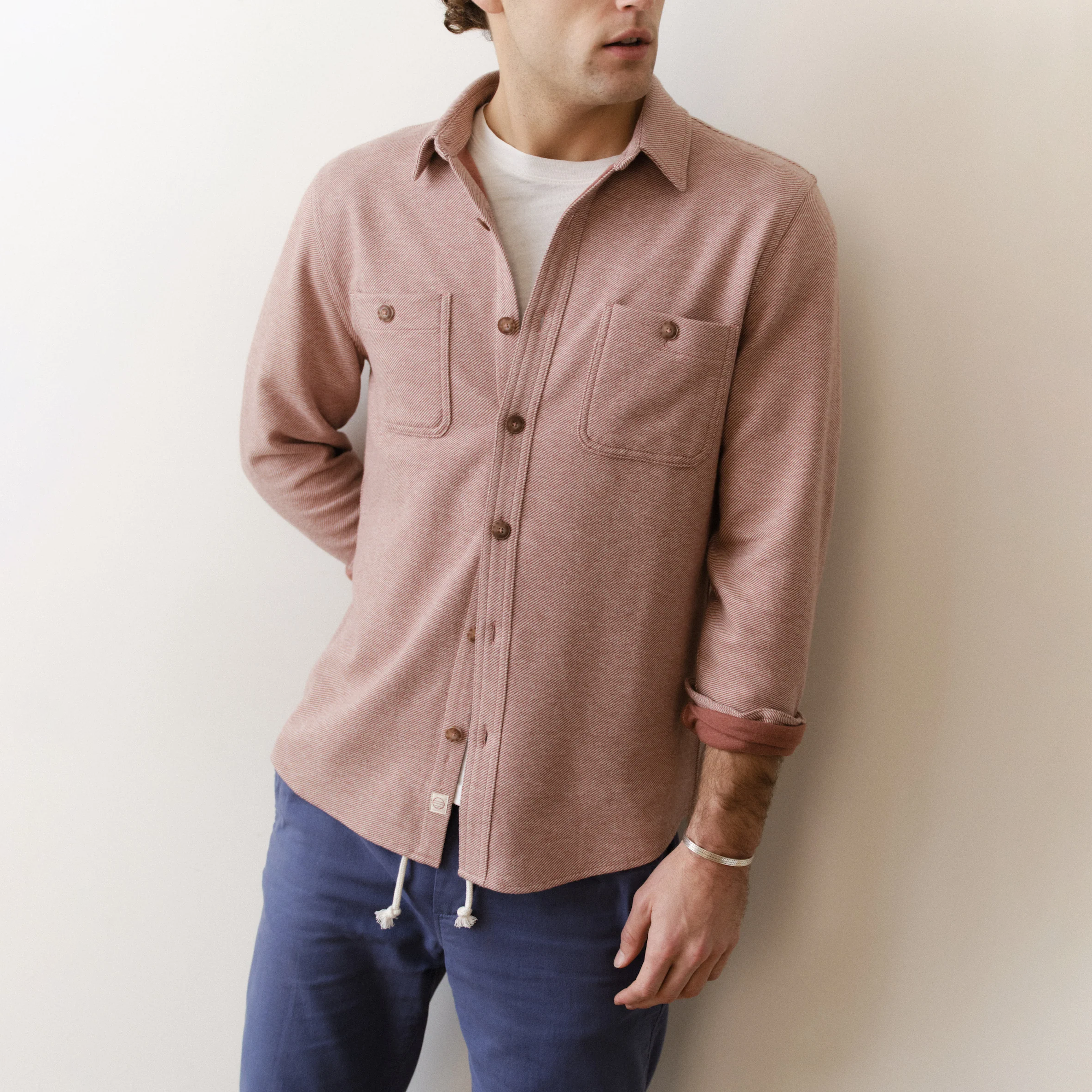 Man wearing a pink button-up shirt and blue pants against a white wall