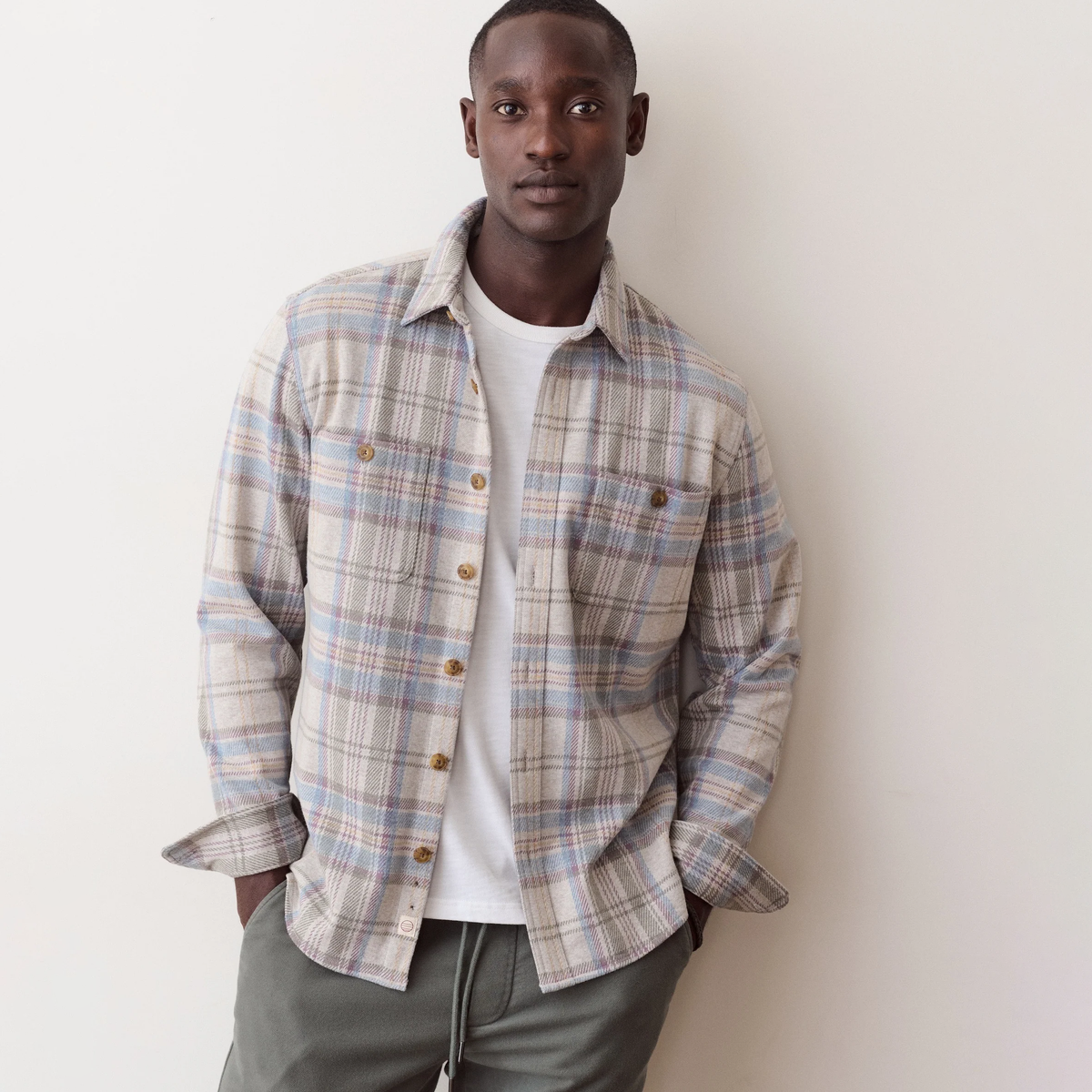 A man wearing a beige, blue, and brown plaid button-down shirt over a white t-shirt and gray sweatpants, standing against a plain off-white wall.