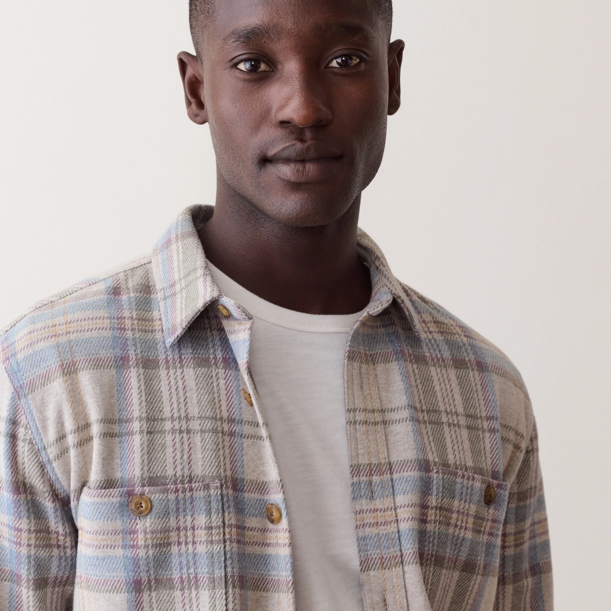 A close-up shot of a man wearing a beige and blue plaid button-up shirt over a white t-shirt, set against a plain off-white background.