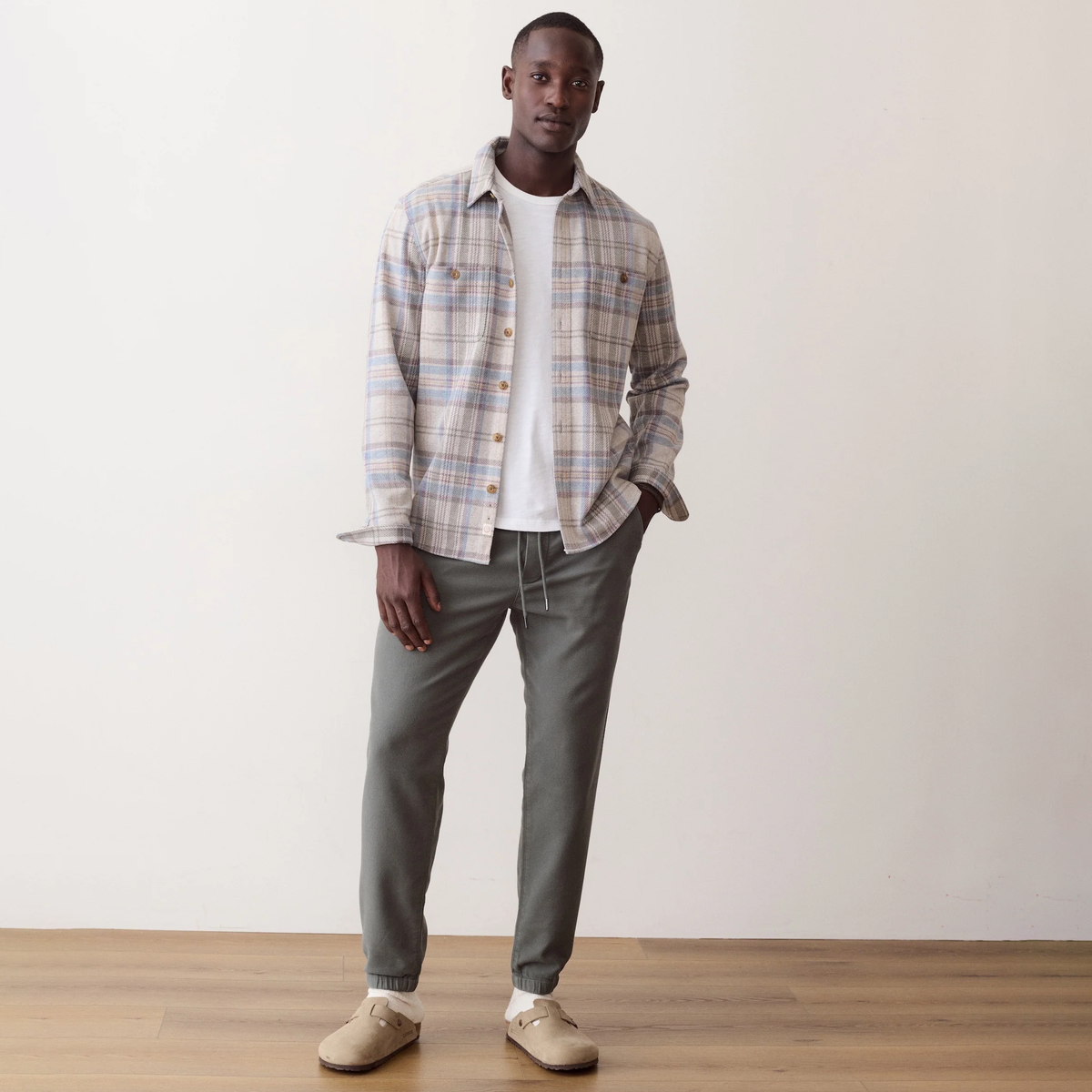 A man stands in a studio wearing a beige and blue plaid button-up shirt over a white t-shirt, paired with olive green drawstring pants and beige slip-on shoes.