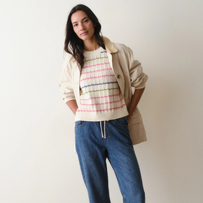 Woman wearing a striped sweater, beige coat, and blue jeans against a plain background