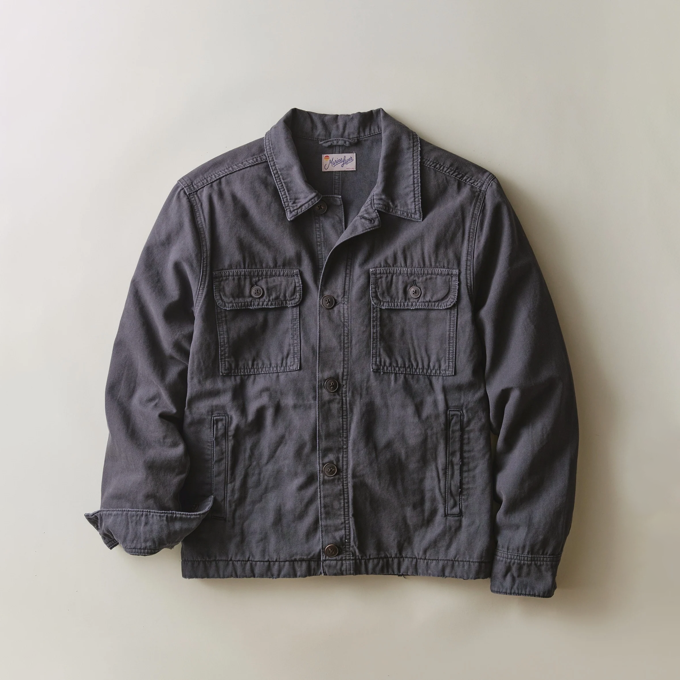 A dark gray canvas utility jacket with a pointed collar, button-up front, and four front pockets, laid flat against a light beige background.