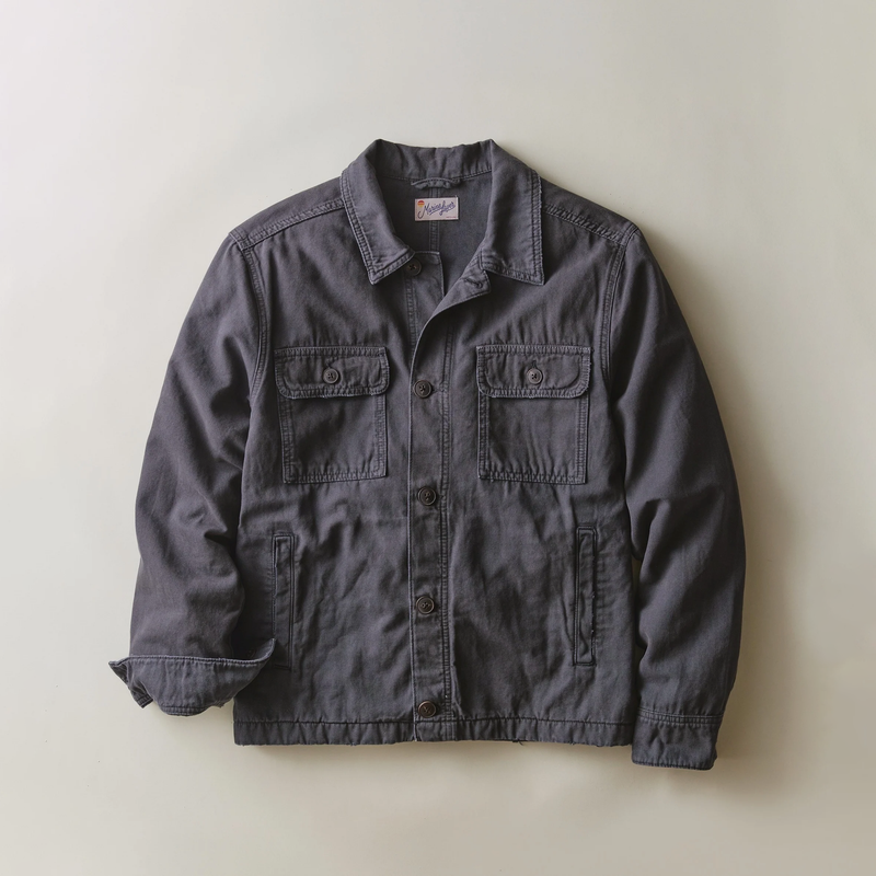 A dark gray canvas utility jacket with a pointed collar, button-up front, and four front pockets, laid flat against a light beige background.