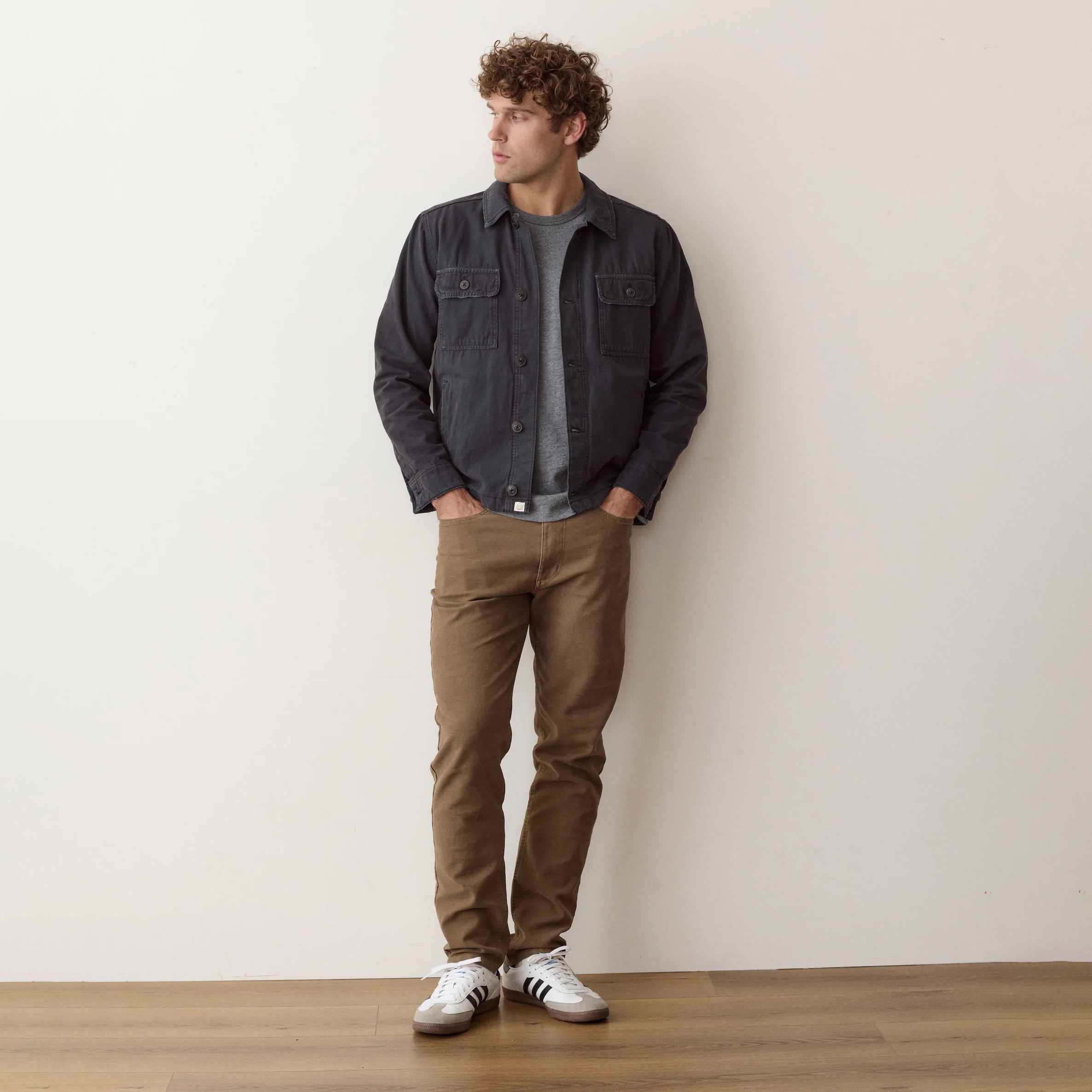 A man stands against a plain off-white wall wearing a dark charcoal gray denim jacket over a gray t-shirt, brown pants, and white sneakers.