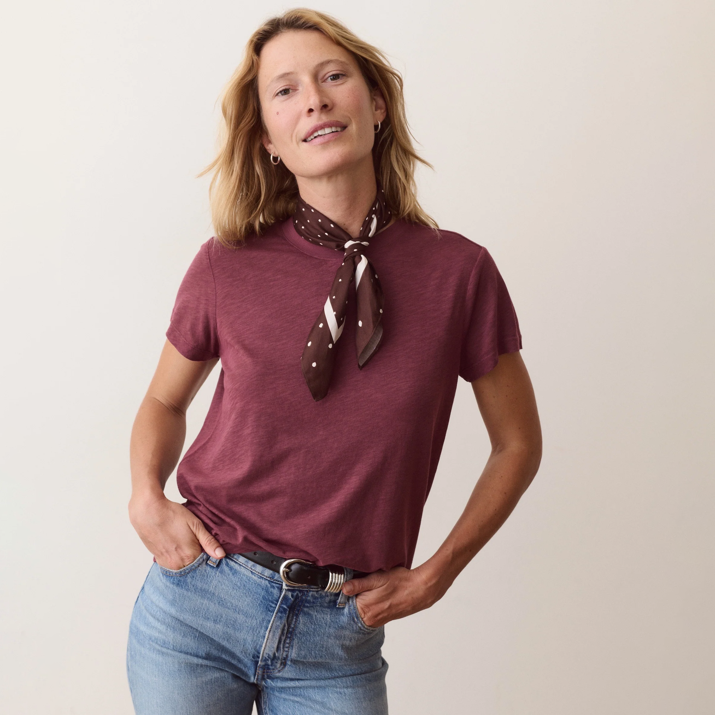 A woman wearing a short-sleeve heathered burgundy t-shirt and a brown patterned neck scarf, paired with blue jeans, standing against a plain light beige background.