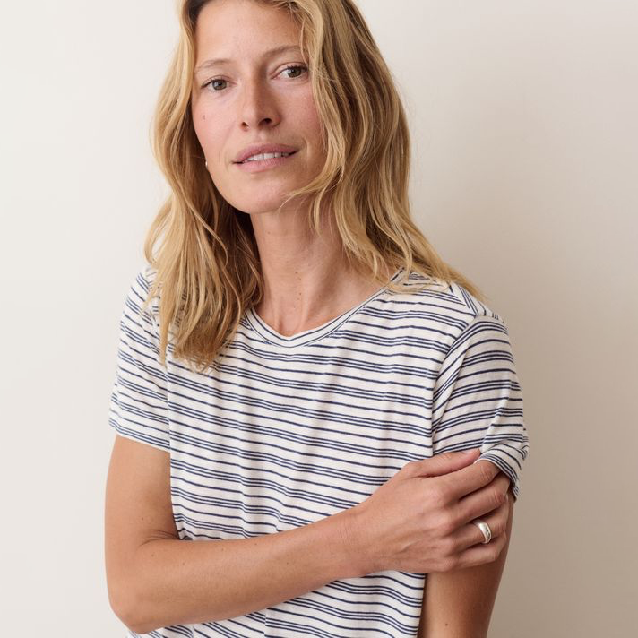 A woman with blonde hair wearing a short-sleeve crewneck t-shirt with thin navy and white horizontal stripes, posed against a plain beige background.