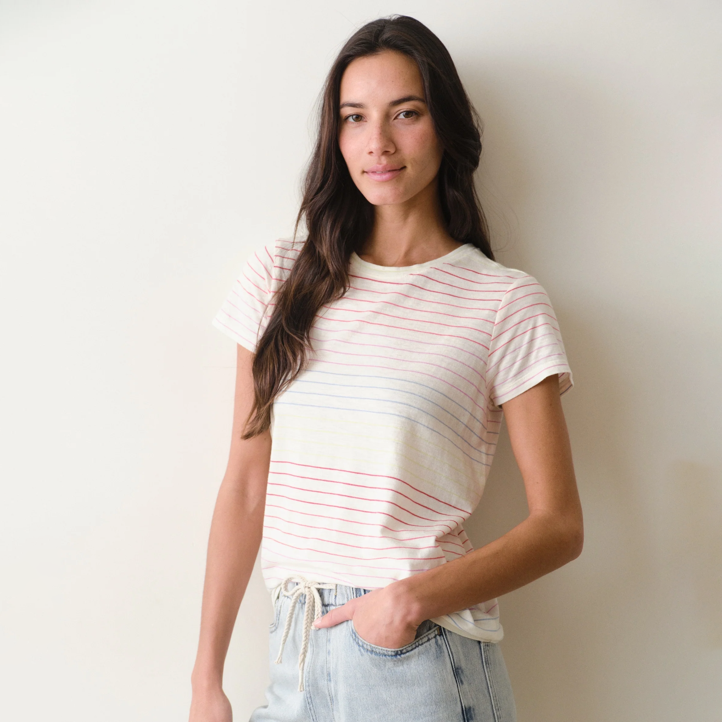 Woman wearing a rainbow stripe tee and jeans on a neutral background.