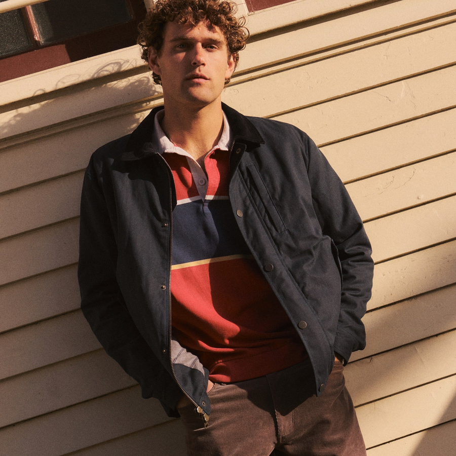 A man wearing a dark navy waxed cotton utility jacket over a red, navy, and white striped polo shirt, leaning against a beige wood-paneled wall.
