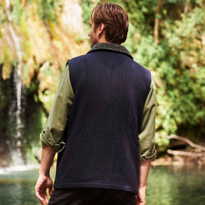 Man wearing a navy vest over a green shirt standing by a forest stream with a waterfall.