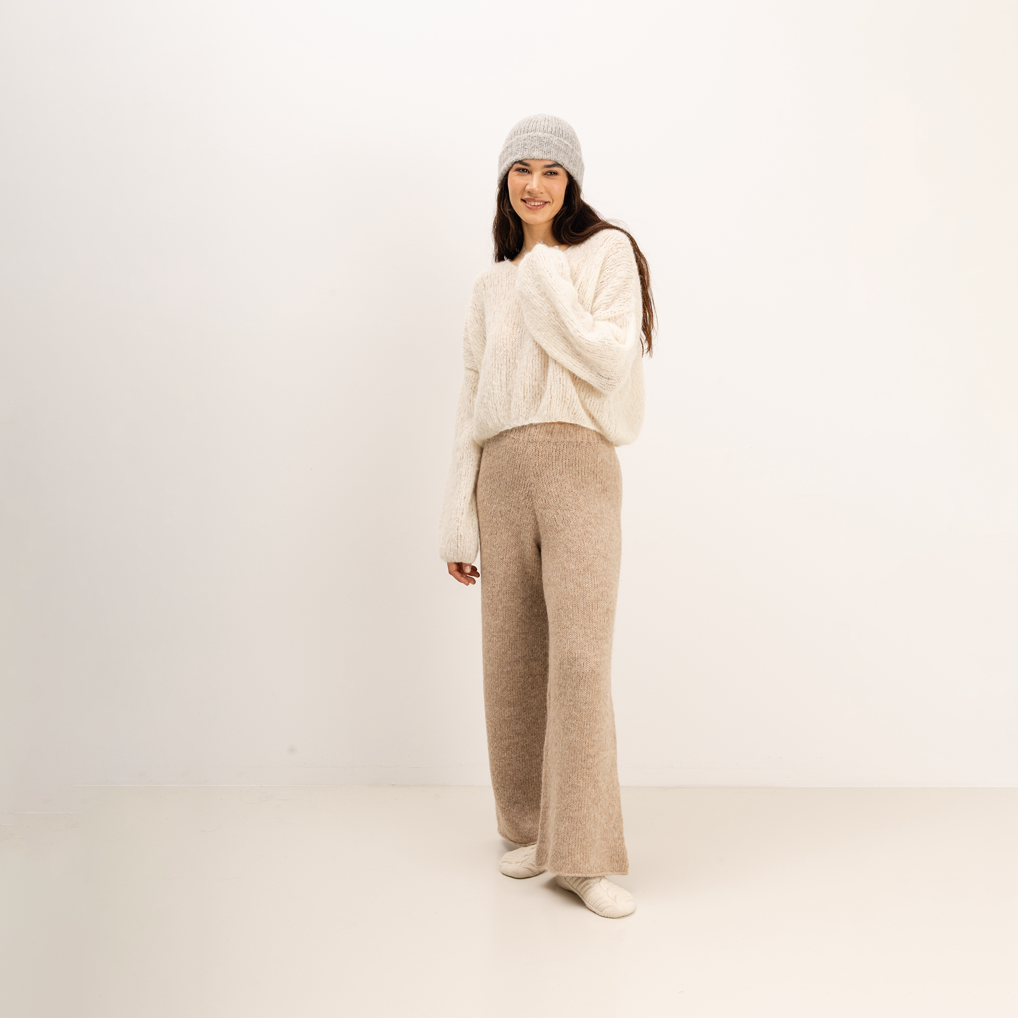 A woman stands in a studio wearing a cream fuzzy knit sweater, beige wide-leg knit pants, a light gray knit beanie, and white shoes against a plain off-white background.