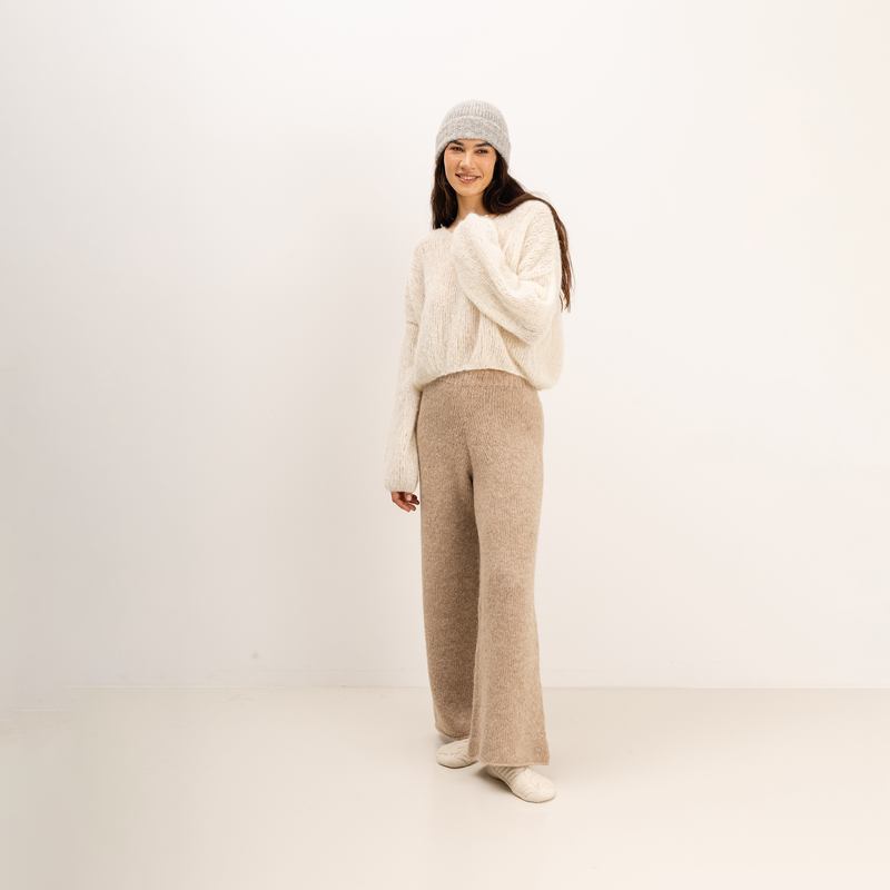 A woman stands in a studio wearing a cream fuzzy knit sweater, beige wide-leg knit pants, a light gray knit beanie, and white shoes against a plain off-white background.