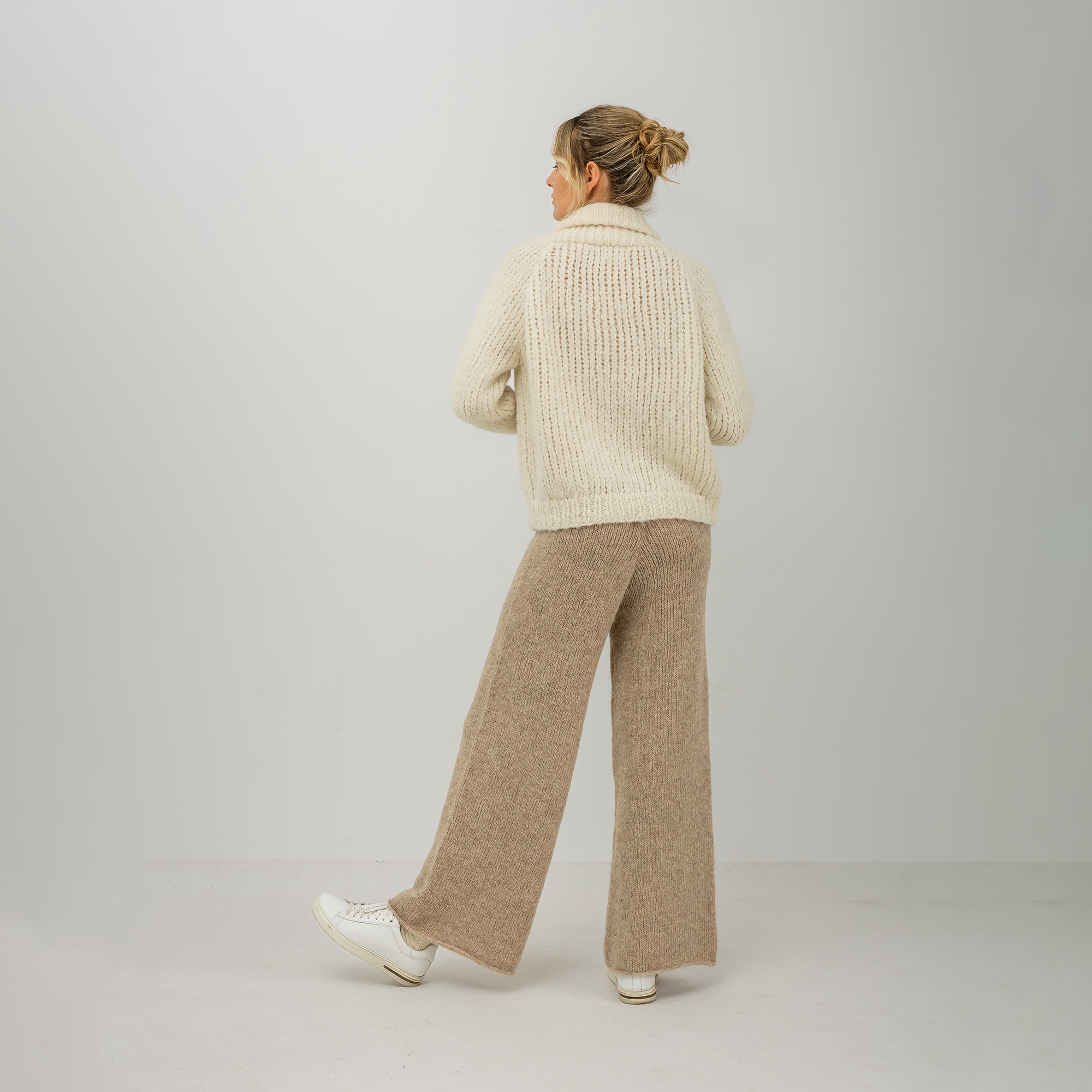 A woman is shown from behind wearing a cream-colored chunky knit sweater and beige wide-leg knit pants, paired with white sneakers, against a plain light gray studio background.