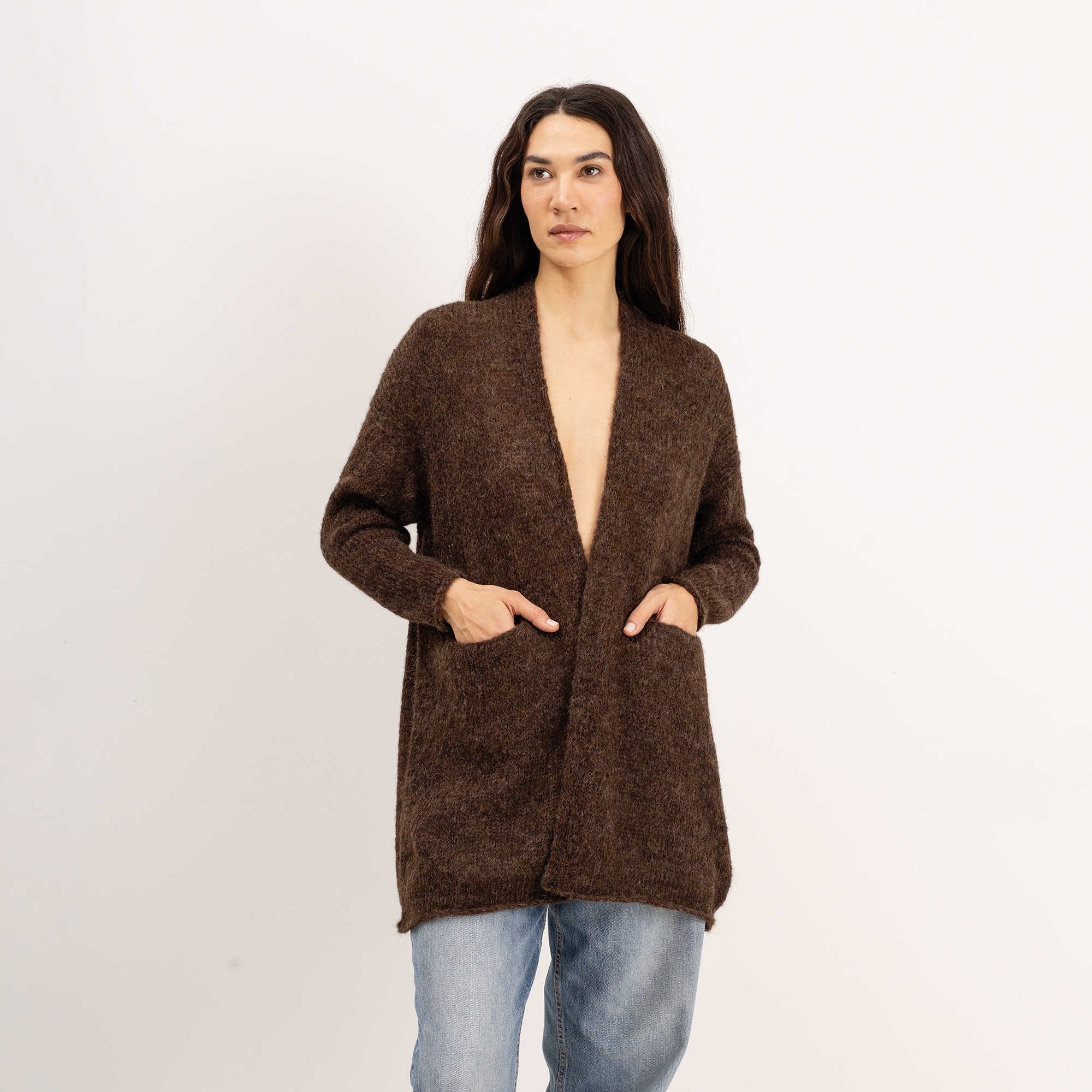 A woman modeling a brown open-front knit cardigan with long sleeves and front pockets, paired with light blue jeans, against a plain white studio background.