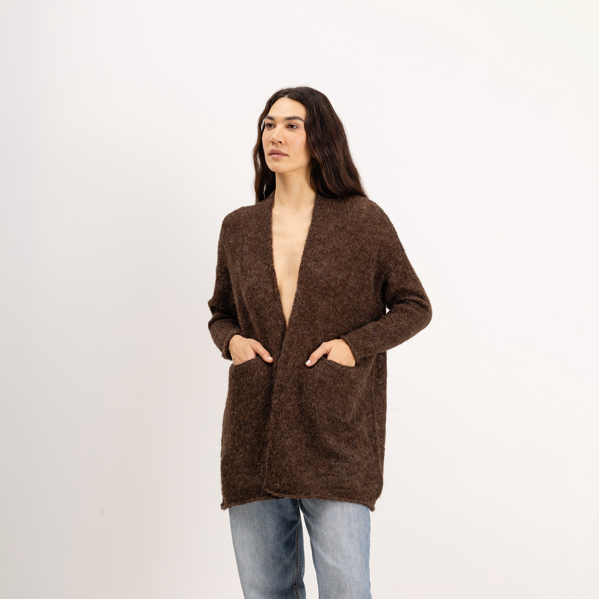 A woman modeling a brown open-front knit cardigan with side pockets, paired with light blue jeans, against a plain white studio background.