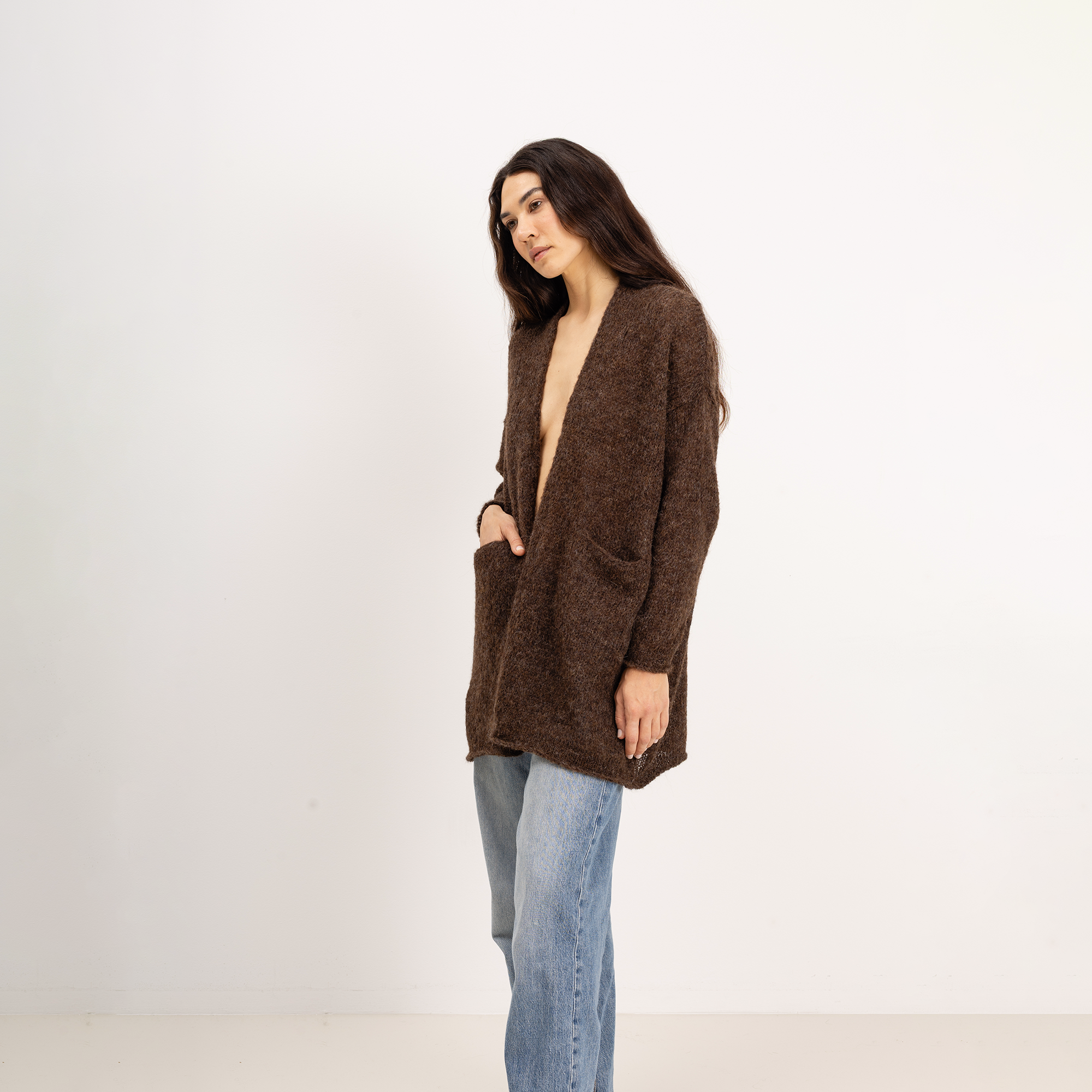 A woman modeling a brown open-front knit cardigan with pockets, paired with light-wash blue jeans, against a plain white studio background.
