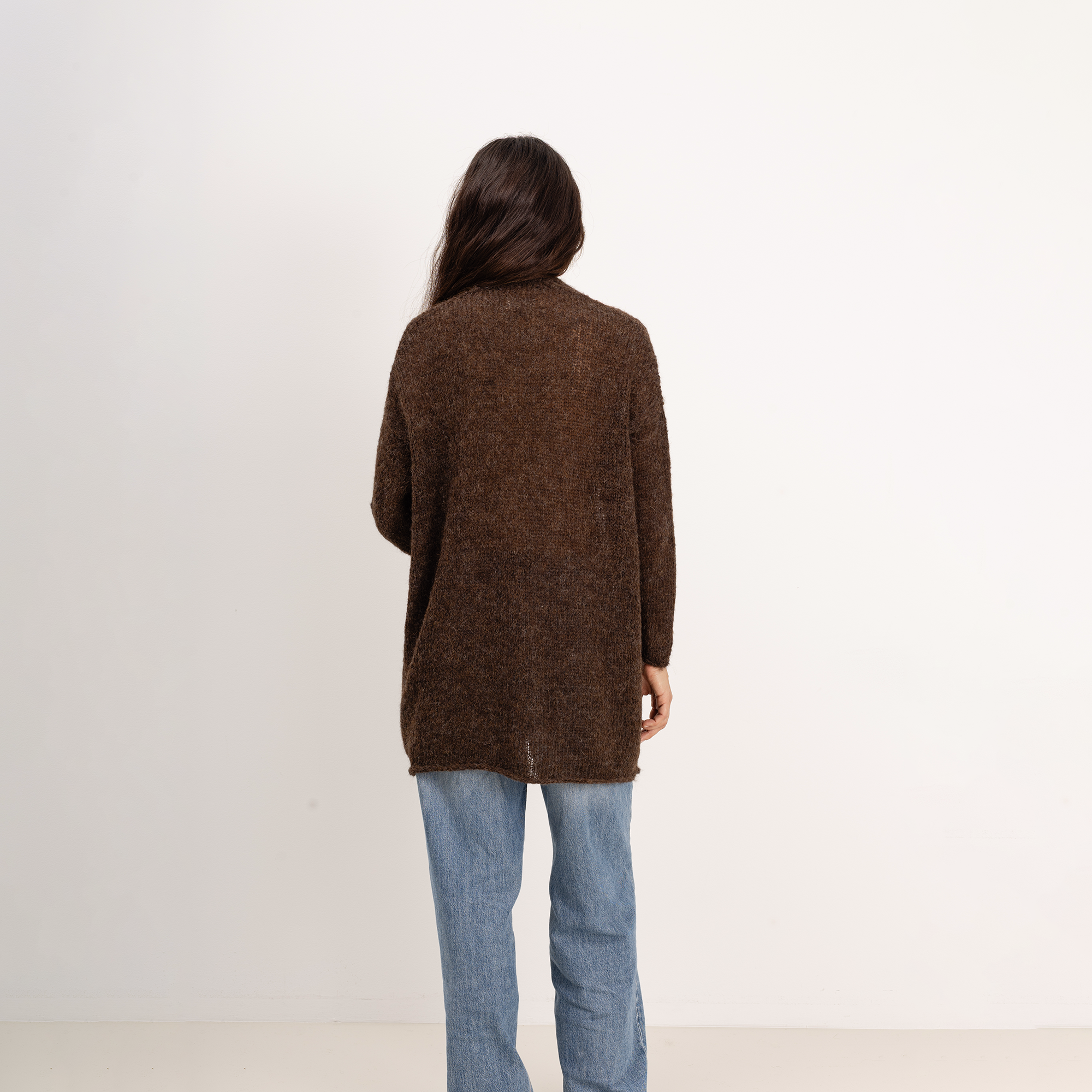 A person viewed from behind wearing a loose, fuzzy brown knit cardigan and light blue jeans against a plain off-white studio background.