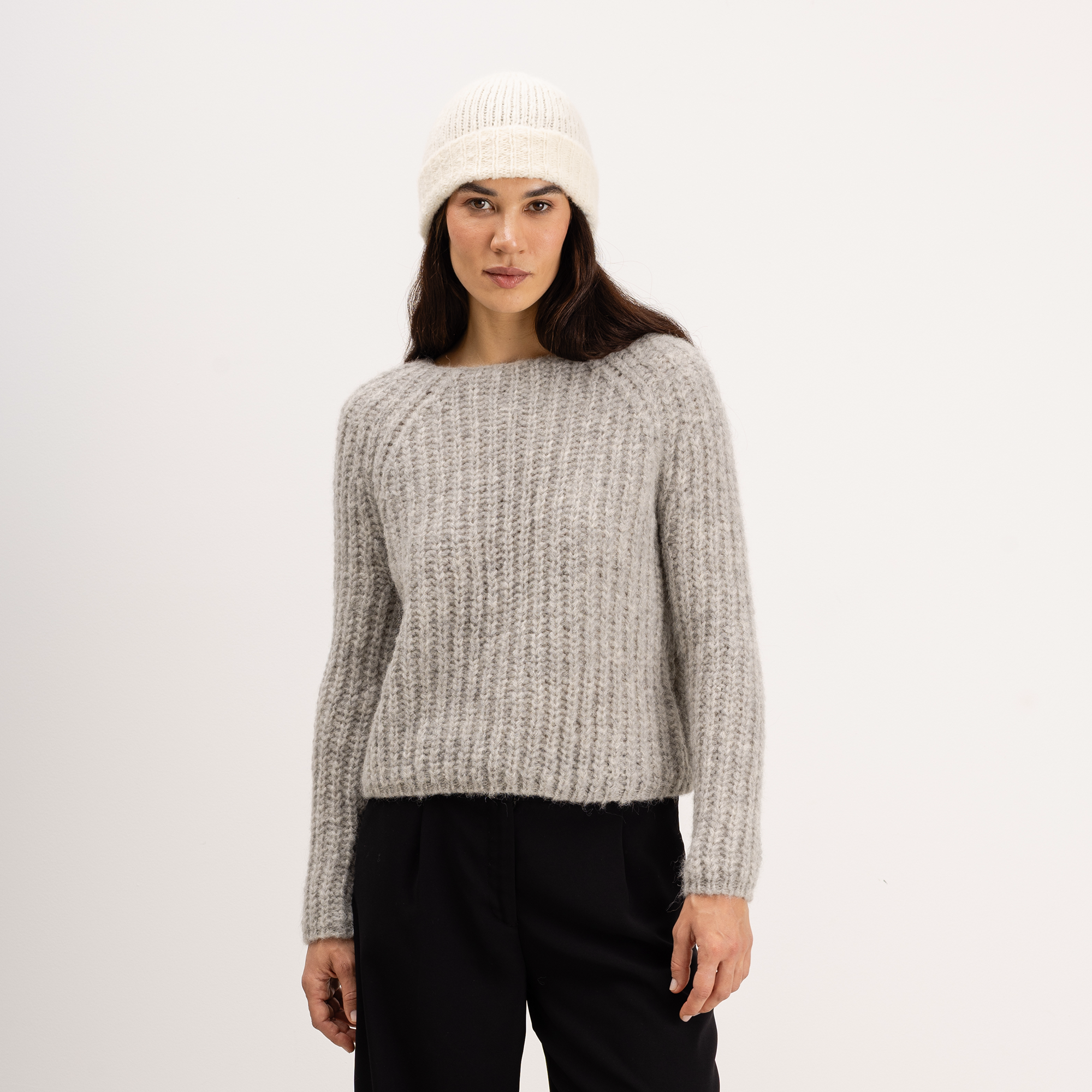 A woman wearing a light gray textured knit sweater, a cream knit beanie, and black pants, standing against a plain white studio background.