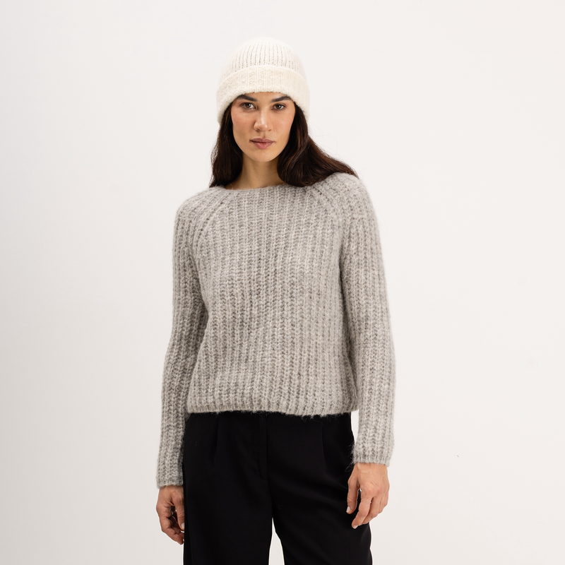 A woman wearing a light gray textured knit sweater, a cream knit beanie, and black pants, standing against a plain white studio background.